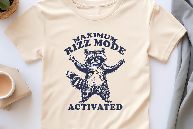 May include: A cream colored t-shirt with a raccoon graphic and the text "Maximum Rizz Mode Activated" in navy blue.