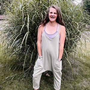 May include: A woman wearing a light green jumpsuit with large pockets and brown shoes. She is standing in front of a large bush.