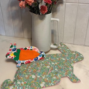 以下が含まれることがあります： ピンクの花が入った白いセラミックピッチャーが、ウサギ型のプレースマットと、ニンジン柄のウサギ型鍋つかみの隣に置かれています。プレースマットはウサギのフローラル柄です。