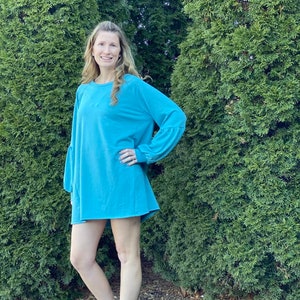 May include: A teal blue long-sleeved dress with a loose fit and a slightly flared hem. The dress has a round neckline and long sleeves with a slight puff at the shoulders.