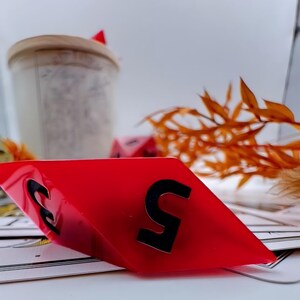 May include: A red, diamond-shaped game piece with the number 5 in black. The piece is angled, revealing the number 3 on the other side. Other game pieces and dried foliage are in the background.