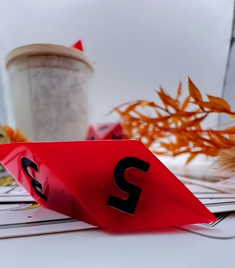 May include: A red, diamond-shaped game piece with the number 5 in black. The piece is angled, revealing the number 3 on the other side. Other game pieces and dried foliage are in the background.