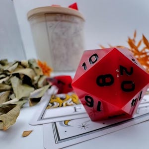 May include: A large, red, twenty-sided die with black numbers, resting on a deck of tarot cards. A candle in a jar and dried leaves are in the background. The die is a prominent feature.