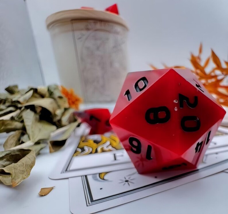 May include: A large, red, twenty-sided die with black numbers, resting on a deck of tarot cards. A candle in a jar and dried leaves are in the background. The die is a prominent feature.