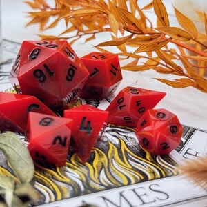 May include: Set of seven red polyhedral dice with black numbering, designed for tabletop role-playing games. The dice include a D4, D6, D8, D10, D12, D20, and a percentile die. The dice are arranged on a printed surface with decorative foliage.