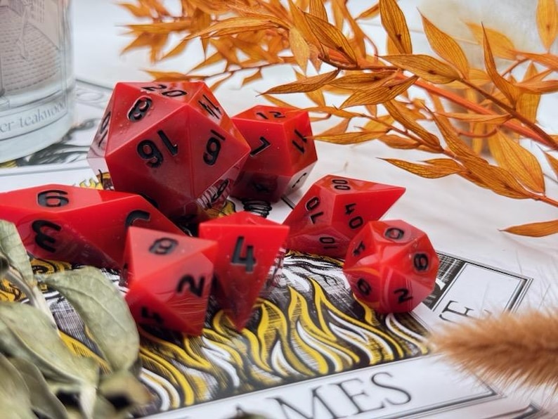May include: Set of seven red polyhedral dice with black numbering, designed for tabletop role-playing games. The dice include a D4, D6, D8, D10, D12, D20, and a percentile die. The dice are arranged on a printed surface with decorative foliage.