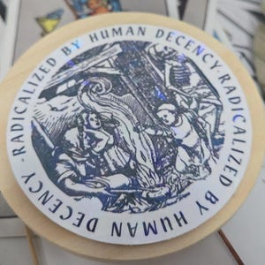 Könnte beinhalten: Ein runder Holzuntersetzer mit einer detaillierten Schwarz-Weiß-Illustration. Der Untersetzer trägt den Text "RADICALIZED BY HUMAN DECENCY" in einem kreisförmigen Muster um das Bild. Das Bild zeigt Figuren und ein Lagerfeuer.