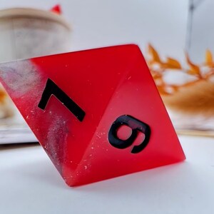May include: A red, resin-cast, polyhedral die with the numbers 7 and 9 in black. The die has a marbled, gray and white effect on one corner. The background is blurred, with a white surface and decorative elements.