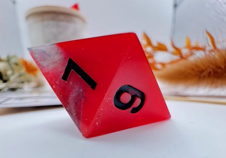 May include: A red, resin-cast, polyhedral die with the numbers 7 and 9 in black. The die has a marbled, gray and white effect on one corner. The background is blurred, with a white surface and decorative elements.