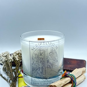 May include: A clear glass candle with a wooden wick and a label that reads "Scriptorium." The label features a black and white illustration of people working. The candle is surrounded by tarot cards, dried flowers, and bundles of wood.