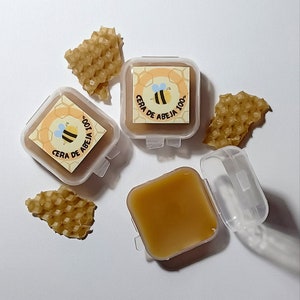Puede incluir: Dos pequeños recipientes de plástico transparente con tapas, cada uno contiene un producto de cera de abeja amarilla. Los recipientes tienen una etiqueta con una abeja de dibujos animados y el texto "Cera de Abeja 100%". Un tercer recipiente está abierto, mostrando la cera de abeja amarilla en el interior. Hay tres trozos de panal en la superficie blanca.