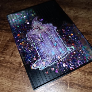 Puede incluir: Una funda rectangular negra con un diseño de vela morada. La vela tiene un efecto de cera goteando. La funda está llena de purpurina colorida, incluyendo morado, azul y verde.