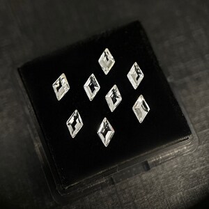 Op de afbeelding: Een zwarte fluwelen doos met acht heldere, gefacetteerde, diamantvormige edelstenen, gerangschikt in een symmetrisch patroon.