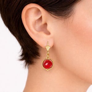 May include: Gold-toned drop earrings featuring a faceted red gemstone. The gemstone is set within a decorative, twisted rope-like frame. The earrings have a lever-back closure and a decorative top.