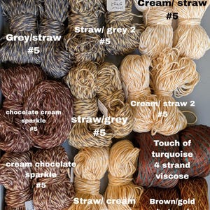 May include: A collection of yarn skeins in various colors, including grey, cream, brown, and gold. Some skeins have a sparkle effect. The image includes text labels such as "Grey/straw #5" and "Touch of turquoise 4 strand viscose."