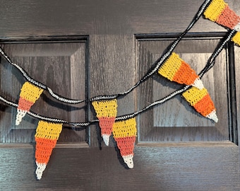May include: A handmade crochet garland featuring candy corn shapes in yellow, orange, and white. The garland is strung on a black cord and is displayed against a dark wooden door. Perfect for Halloween or fall decor.