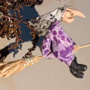 May include: A hanging ornament depicting a witch figure with a pale face and hooked nose, wearing a white headscarf with black floral patterns and a purple dress with a darker purple abstract print, riding a straw broom with a floral fabric wrap.