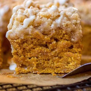 May include: A close-up of a square pumpkin spice cake with a crumb topping and white icing drizzle. The cake is sitting on a wire rack and a piece of parchment paper. The image is well-lit and shows the texture of the cake.