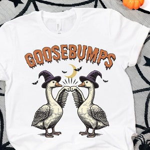 May include: White t-shirt with a graphic of two geese wearing witch hats and the text "GOOSEBUMPS" in orange dripping letters. The geese are fist bumping each other.