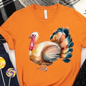 Puede incluir: Camiseta naranja con una ilustración de acuarela de un pavo. El pavo es marrón, beige y azul con una barba rosa.