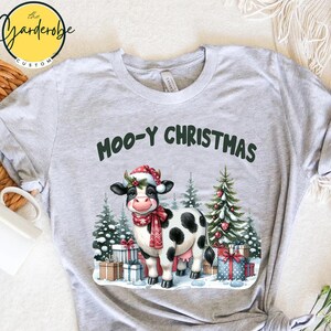 Peut inclure: T-shirt gris clair avec l'inscription "Moo-y Christmas" en vert. Le t-shirt présente une vache de dessin animé portant un bonnet et une écharpe de Père Noël, entourée de sapins et de cadeaux de Noël. Le t-shirt a des manches retroussées.