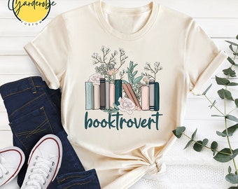 Może przedstawiać: Beżowa koszulka z nadrukiem stosu książek z kwiatami i tekstem "booktrovert".