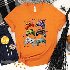 Op de afbeelding: Oranje T-shirt met een afbeelding van zes videogamecontrollers versierd met Halloween-thema's, waaronder pompoenen, vleermuizen en een heksenhoed.