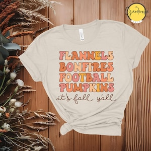 Könnte beinhalten: Cremefarbenes T-Shirt mit dem Text "FLANNELS BONFIRES FOOTBALL PUMPKINS it's fall y'all" in einem Retro-Schriftstil. Der Text ist in Orange- und Rosatönen gehalten. Das Shirt ist an der Taille geknotet.