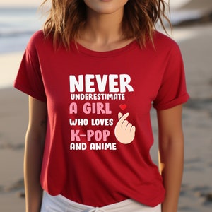 May include: Red t-shirt with the text "Never underestimate a girl who loves K-pop and anime" in white. A pink heart and a pink hand gesture are included in the design.