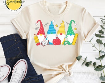 May include: A beige t-shirt featuring a design of four gnomes wearing colourful hats and holding school supplies. The gnomes are holding a pencil, a book, an apple, and a ruler. The text "the garde robe custom" is in the top left corner of the shirt.