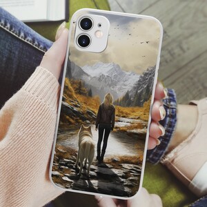 May include: A white phone case with a scenic image of a woman and a dog walking through a snowy mountain landscape. The case has a camera cutout and a white background.