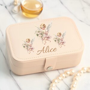 May include: A blush pink rectangular jewelry box with a clasp closure. The lid features three cherub illustrations and the name "Alice" in gold script. A pearl necklace is visible in the foreground.