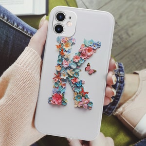May include: White phone case with a floral design in the shape of the letter K. The flowers are pink, blue, and yellow. There is a small butterfly on the case.