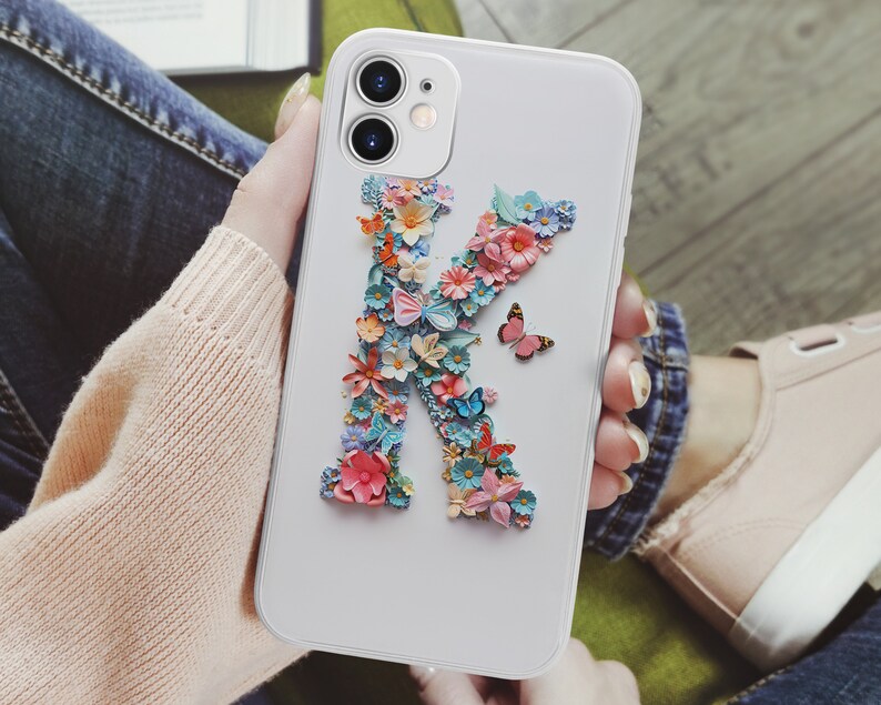 May include: White phone case with a floral design in the shape of the letter K. The flowers are pink, blue, and yellow. There is a small butterfly on the case.