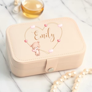 May include: A light pink rectangular jewelry box with a clasp closure. The lid features a gold heart design with the name "Emily" in cursive, surrounded by pink hearts and a teddy bear illustration.