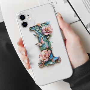 May include: A white iPhone case with a 3D floral design featuring a large letter "I" with butterflies and pink flowers. The case has a camera cutout and a white background.
