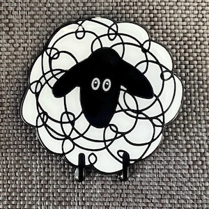 May include: A decorative sheep ornament with a black face and legs, and a white body outlined in black. The body is formed from looping lines. The sheep has two small, round eyes. A fun, quirky design.