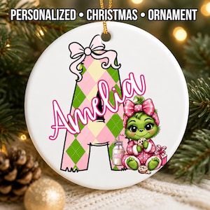 May include: A round white ceramic Christmas ornament featuring a large letter A with a pink and green argyle pattern, topped with a pink bow. The name Amelia is written in pink script. A green character sits nearby with a cookie and milk.