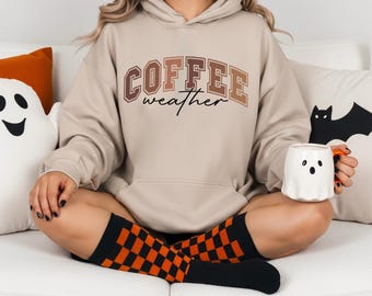 Puede incluir: Una sudadera con capucha beige con el texto "COFFEE weather" en letras marrones y beige. La persona lleva calcetines a cuadros negros y naranjas y sostiene una taza blanca con forma de fantasma con un asa naranja. Hay cojines con temática de Halloween en el fondo.