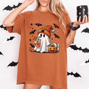 Puede incluir: Camiseta de color naranja quemado con un diseño de Halloween. El diseño presenta un fantasma con gorro y bebida, rodeado de murciélagos, estrellas y una calabaza. La camiseta tiene mangas cortas y un corte holgado.