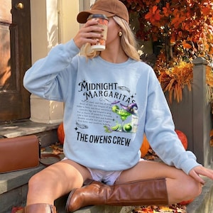 Könnte beinhalten: Ein hellblaues Langarm-Sweatshirt mit einem Grafikdruck, der den Text Midnight Margaritas und The Owens Crew sowie eine Abbildung eines Margaritaglases, eines Hexenhutes und eines Zaubertrankrezepts in schwarzer Schrift zeigt.