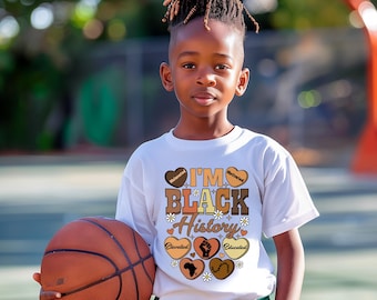 Op de afbeelding: Een jongere draagt een wit t-shirt met de tekst "I'M BLACK History" in bruine en oranje letters, met een bruine basketbal in de hand. Het shirt heeft hart- en bloemafbeeldingen. De persoon draagt een groene korte broek.