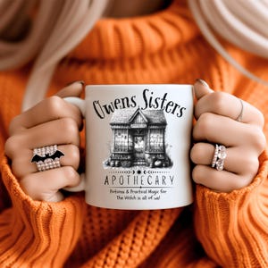 May include: White ceramic mug with the text "Owens Sisters Apothecary" and a detailed illustration of a shop. The mug also includes the phrase "Potions & Practical Magic for The Witch in all of us!" in black lettering.