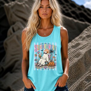 May include: A light blue sleeveless tank top featuring a graphic print of a ghost wearing sunglasses and sitting in a beach chair. The design includes a pumpkin, palm tree, tropical fish, and the text "SUMMERWEEN" with "SPOOKY DAYS SALTY WAVES".