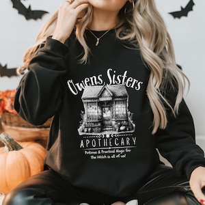 Könnte beinhalten: Ein schwarzes Langarm-Sweatshirt mit einem weißen Grafikdruck eines Gebäudes mit der Aufschrift Apothecary. Der Text lautet Owens Sisters Apothecary, Potions & Practical Magic for The Witch in all of us! mit einer detaillierten Ladenillustration.
