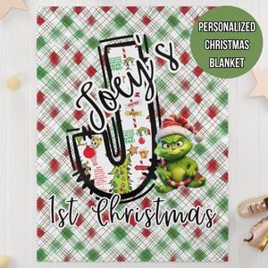 May include: A personalized Christmas blanket with a green and red plaid pattern. The blanket features the name "Joey's" in a stylized font, along with a Grinch character wearing a Santa hat. The text "1st Christmas" is also visible.