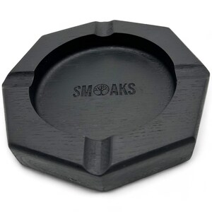 Könnte beinhalten: Schwarzer achteckiger Aschenbecher aus Holz mit einem Baumlogo und dem Text "SMOAKS" auf der Unterseite.