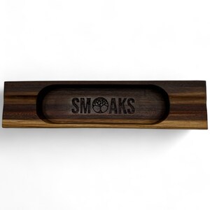 Könnte beinhalten: Ein Holz-Rolltablett mit dunkelbrauner Oberfläche und erhöhtem Rand. Das Tablett hat eine kleine, kreisförmige Vertiefung in der Mitte mit dem Text "SMOAKS" und einem eingravierten Baumlogo.