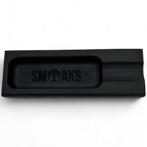 Könnte beinhalten: Ein rechteckiger, schwarzer Aschenbecher mit einer vertieften Fläche und einer gebogenen Kante. Das Wort "SMOAKS" ist in der Mitte eingraviert, mit einem Baumlogo. Der Aschenbecher besteht aus dunklem Material.