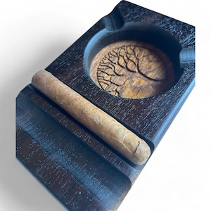 Könnte beinhalten: Ein dunkler Aschenbecher aus Holz mit einem Lebensbaum-Design und einer integrierten Zigarrenstütze.